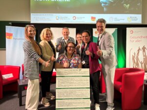 Gruppe von Menschen bei einer Gesundheitskonferenz in Österreich, lächeln und Daumen hoch, vor einem Gesundheitsplakat.