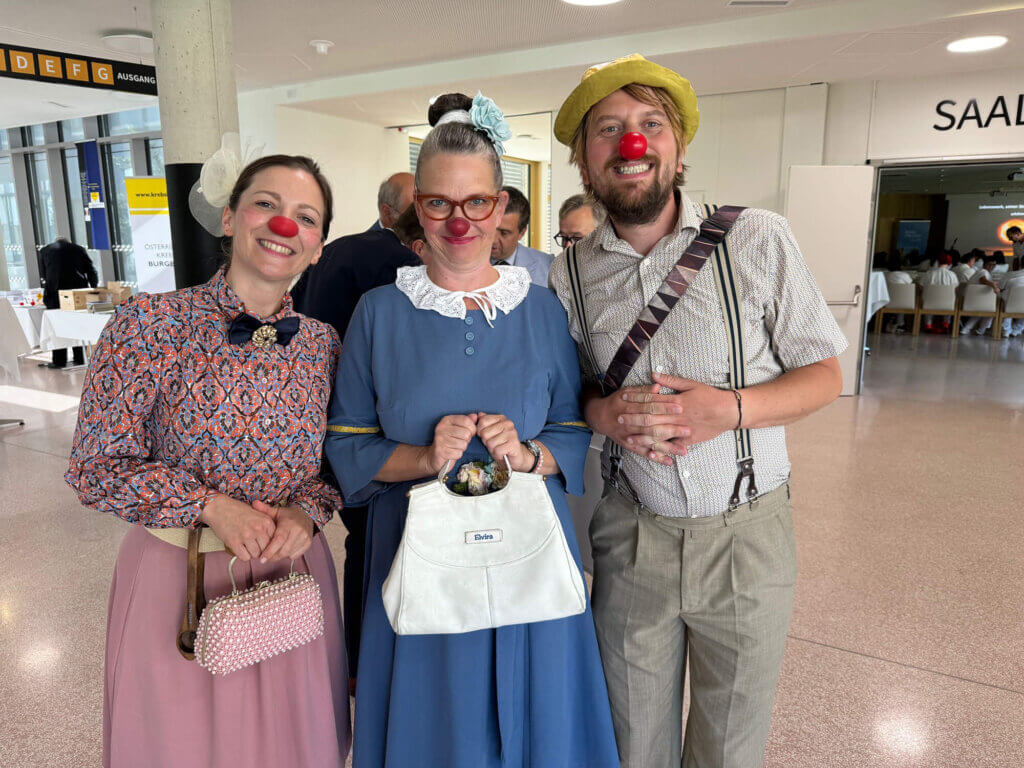 Drei fröhliche Menschen in Clownskostümen mit roten Nasen lächeln bei einer Veranstaltung in einem Saal.