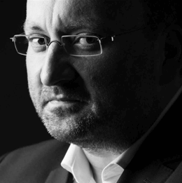 Schwarz-Weiß-Porträt eines ernsten Mannes mit Brille und Bart im Anzug, halbseitiges Profil.