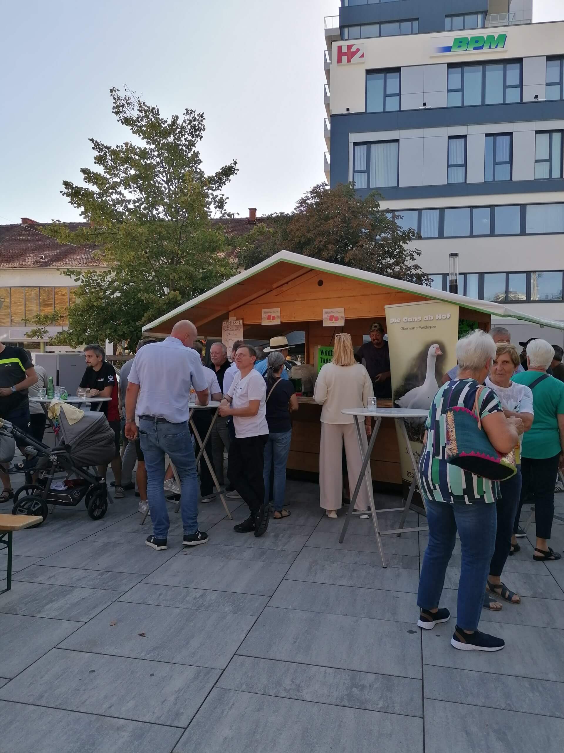 Leute versammeln sich bei einem Außenstand auf einem Marktplatz, modernes Gebäude im Hintergrund.