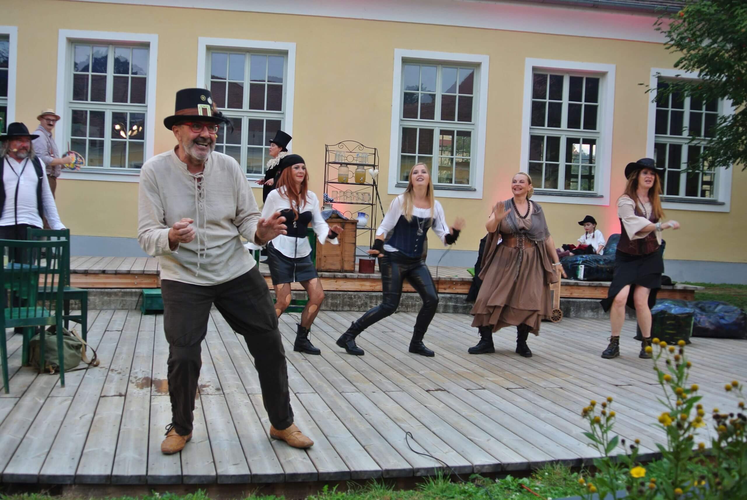 Gruppe in historischen Kostümen tanzt fröhlich auf einer Holzterrasse bei einem Freilufttheaterfest.