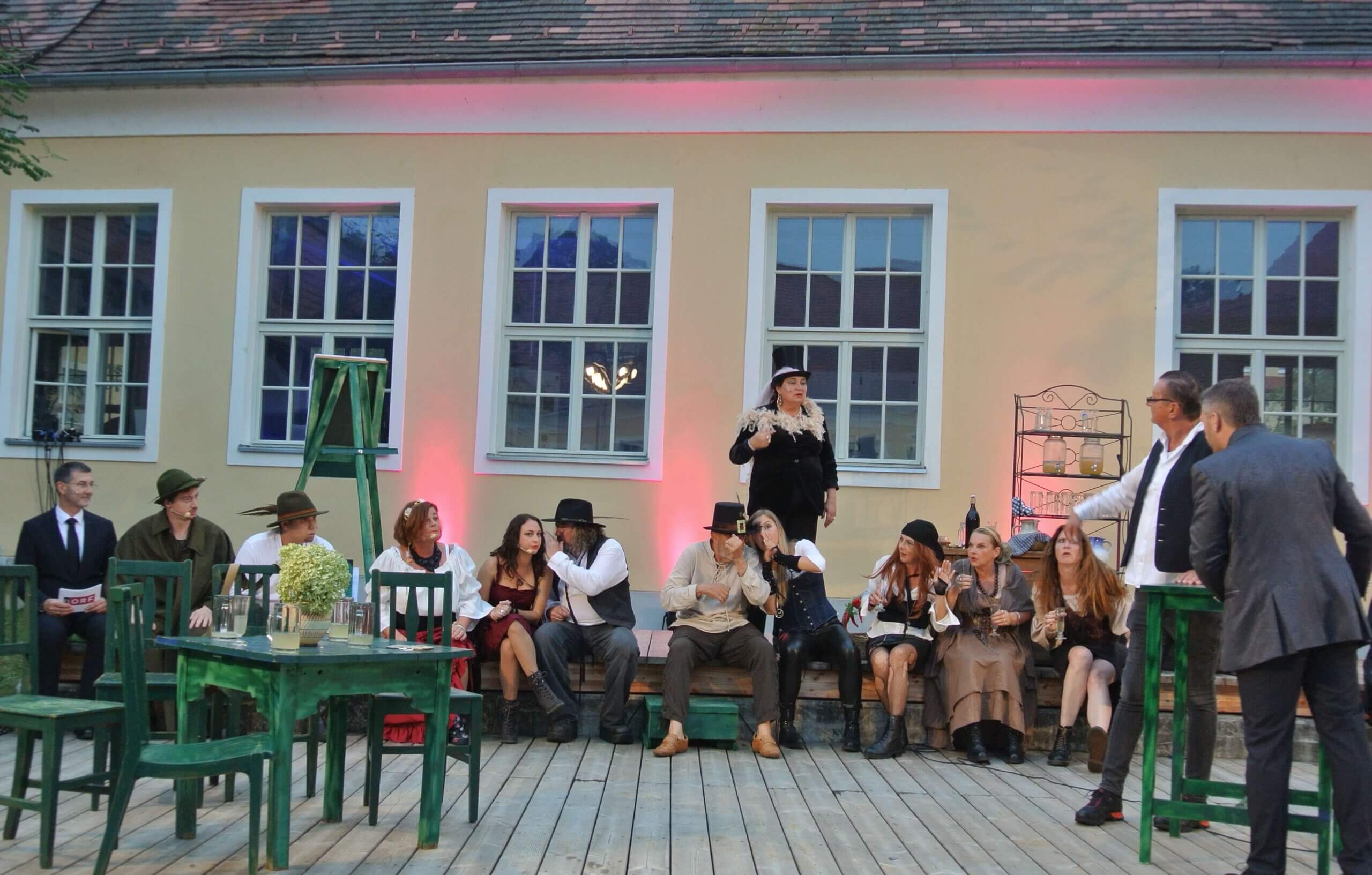 Theateraufführung im Freien, kostümierte Darsteller auf Bühne vor gelbem Gebäude mit beleuchteten Fenstern.