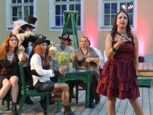 Theaterszene mit kostümierten Schauspielern bei einer lebhaften Aufführung im Freien vor farbig beleuchteter Kulisse.