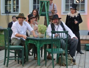 Gruppe fröhlicher Menschen in Vintage-Kleidung bei einem Outdoor-Event, lächelnd an einem Tisch mit Getränken.