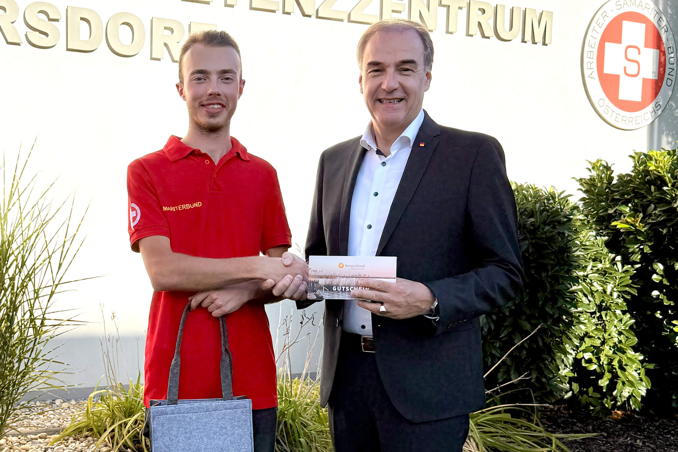 Zwei Männer schütteln Hände vor einem Gebäude des Arbeiter-Samariter-Bunds Österreich, einer hält einen Gutschein. Nico Müller ist Zivildiener des Jahres.