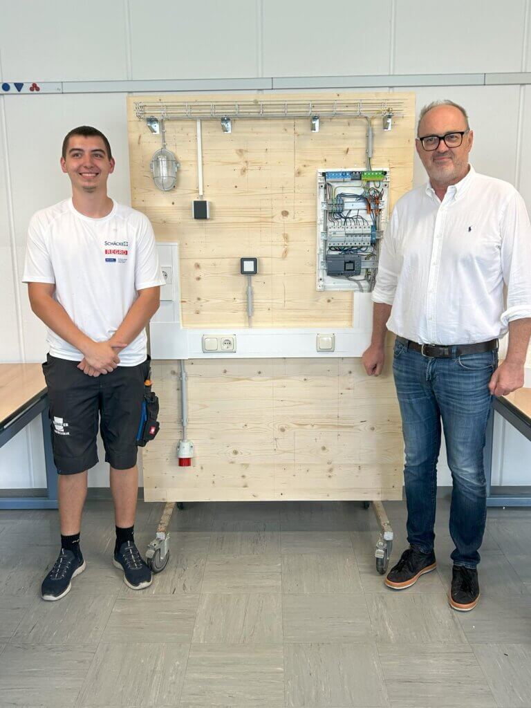 Zwei Männer vor einer elektrischen Schalttafel, Darstellung einer Elektroinstallation in einem Seminarraum.
