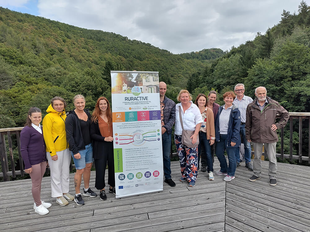 Gruppe von Menschen posiert neben RURACTIVE-Plakat auf einer Holzterrasse mit Wald im Hintergrund.