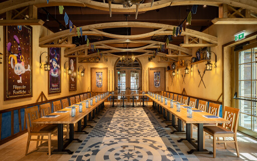 Gemütlicher, rustikaler Gastraum mit langen Holztischen und Wanddekoration im Stil eines mittelalterlichen Festsaals.