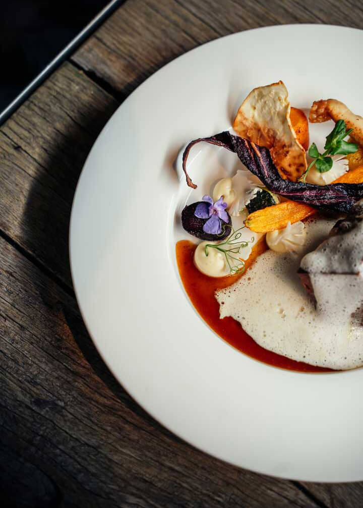 Gourmetgericht auf weißem Teller mit Schaum, Gemüsechips und essbaren Blumen auf rustikalem Holztisch.
