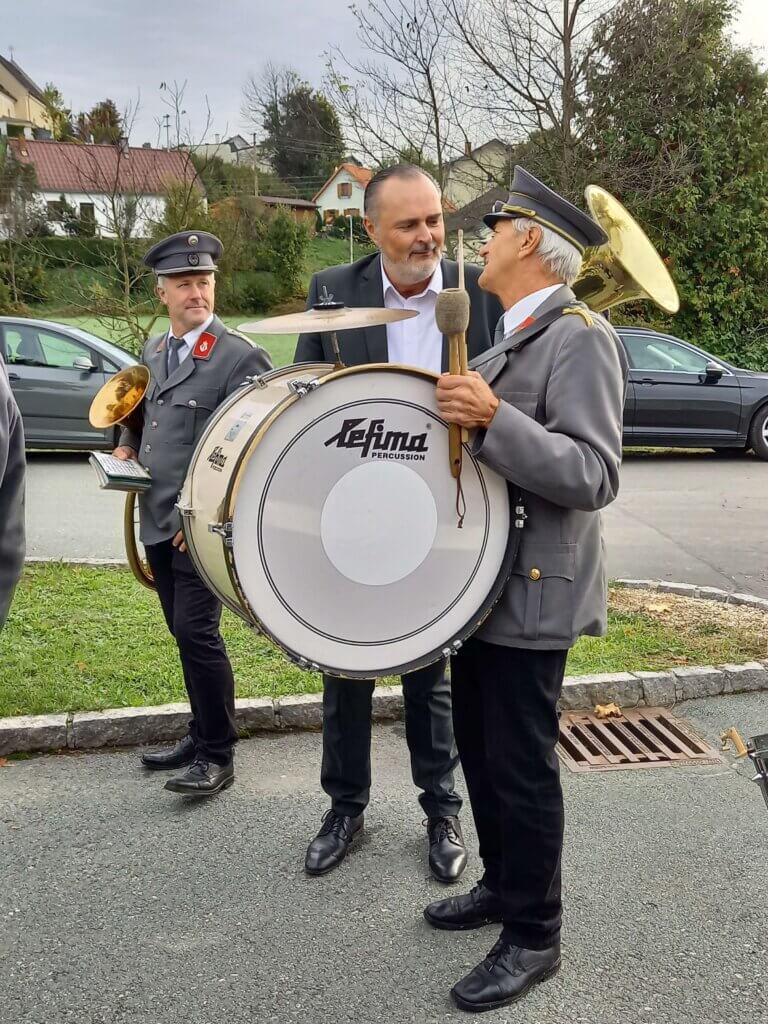 Musikkapelle in Uniformen beim Gespräch an Straßenrand, ländliche Kulisse. Aufnahme für regionales Magazin Prima.