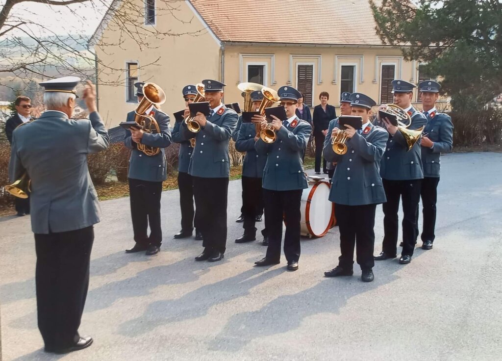 Blaskapelle in traditionellen Uniformen spielt im Freien, dirigiert von einem Kapellmeister vor einem historischen Gebäude.