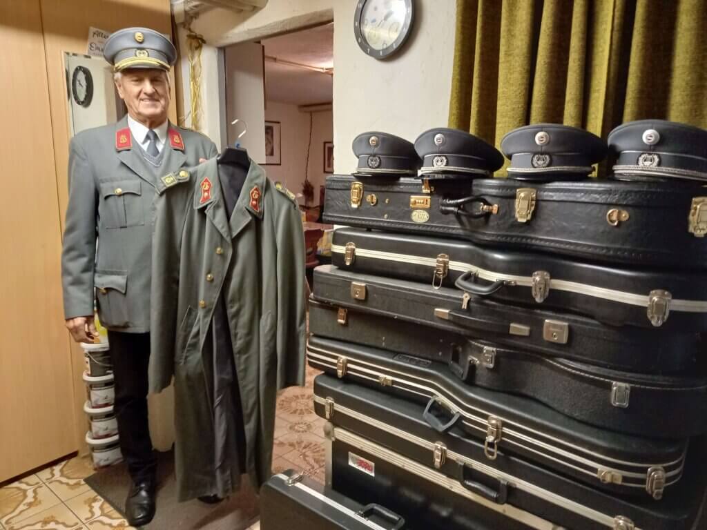 Ein Mann in Uniform zeigt stolz eine weitere Jacke, neben ihm Koffer mit Uniformmützen, im Fokus einer Sammlung.