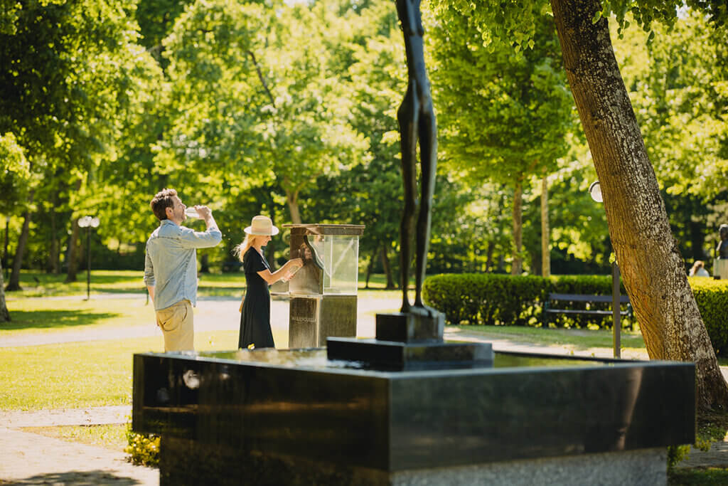 Paar genießt Wasser aus einem Brunnen im sonnigen Park, umgeben von grünen Bäumen, vermittelt entspannte Atmosphäre.