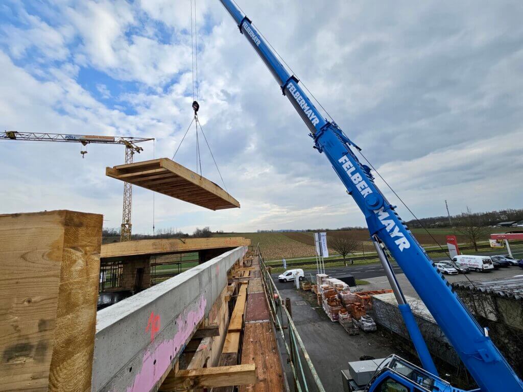 3a5d9ce5-9802-423e-9dc4-cb6c54dc7c2a©PIK SÜD Gmbh Kran hebt Holzplatte auf Baustelle. Regionales Bauprojekt mit Blick auf die umliegende Landschaft. Foto für Prima Magazin.