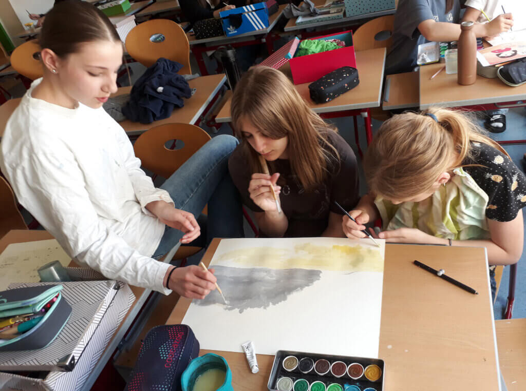 Schülerinnen malen gemeinsam im Kunstunterricht an einem Tisch. Kreatives Arbeiten in der Schule, inspiriert durch Prima Magazin.