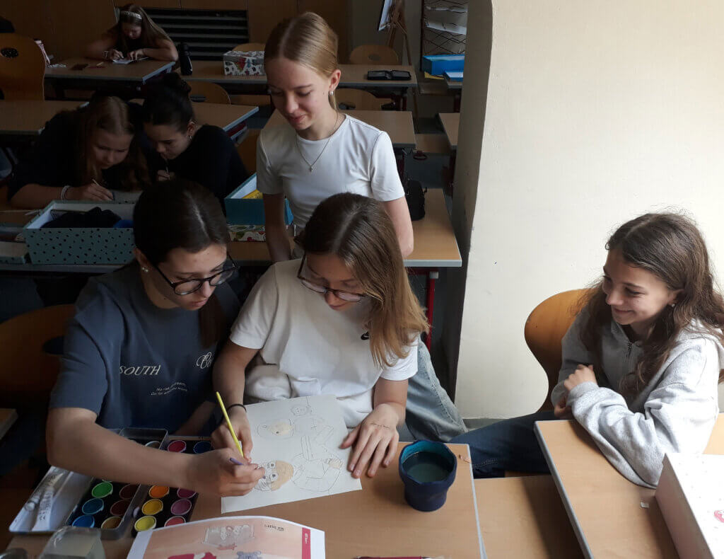 Schülerinnen in einem Klassenraum arbeiten gemeinsam an einem Kunstprojekt und nutzen Aquarellfarben, lächelnd und konzentriert.