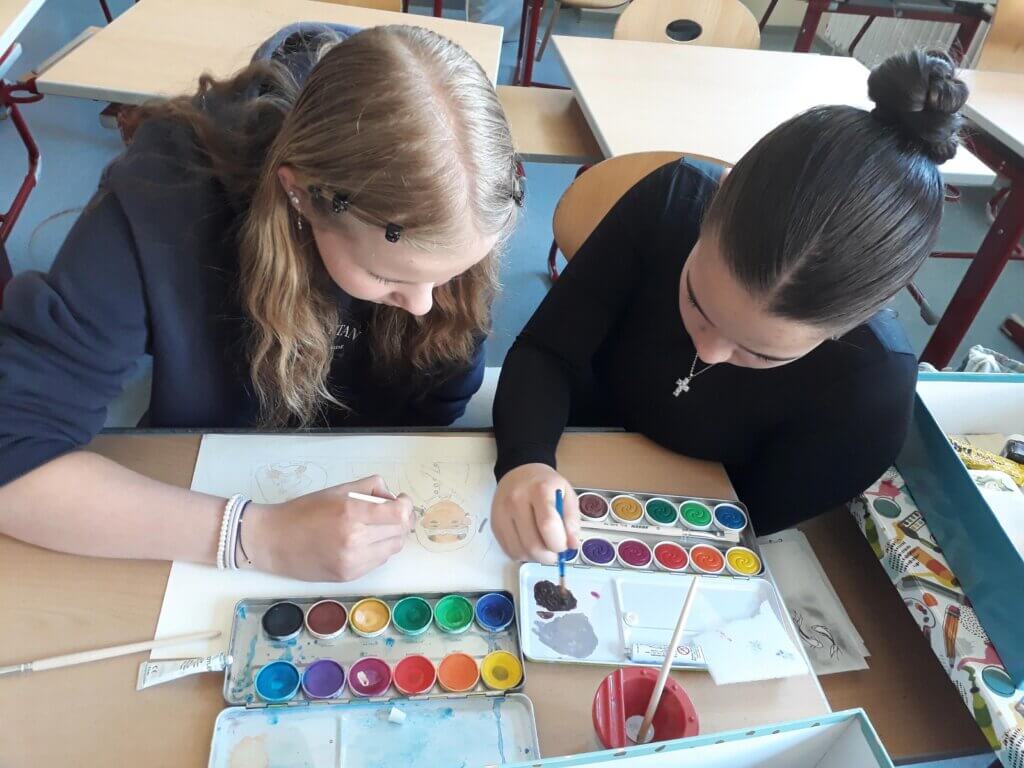 Zwei Schülerinnen malen im Kunstunterricht mit Wasserfarben an einem Holztisch in einem Klassenzimmer.