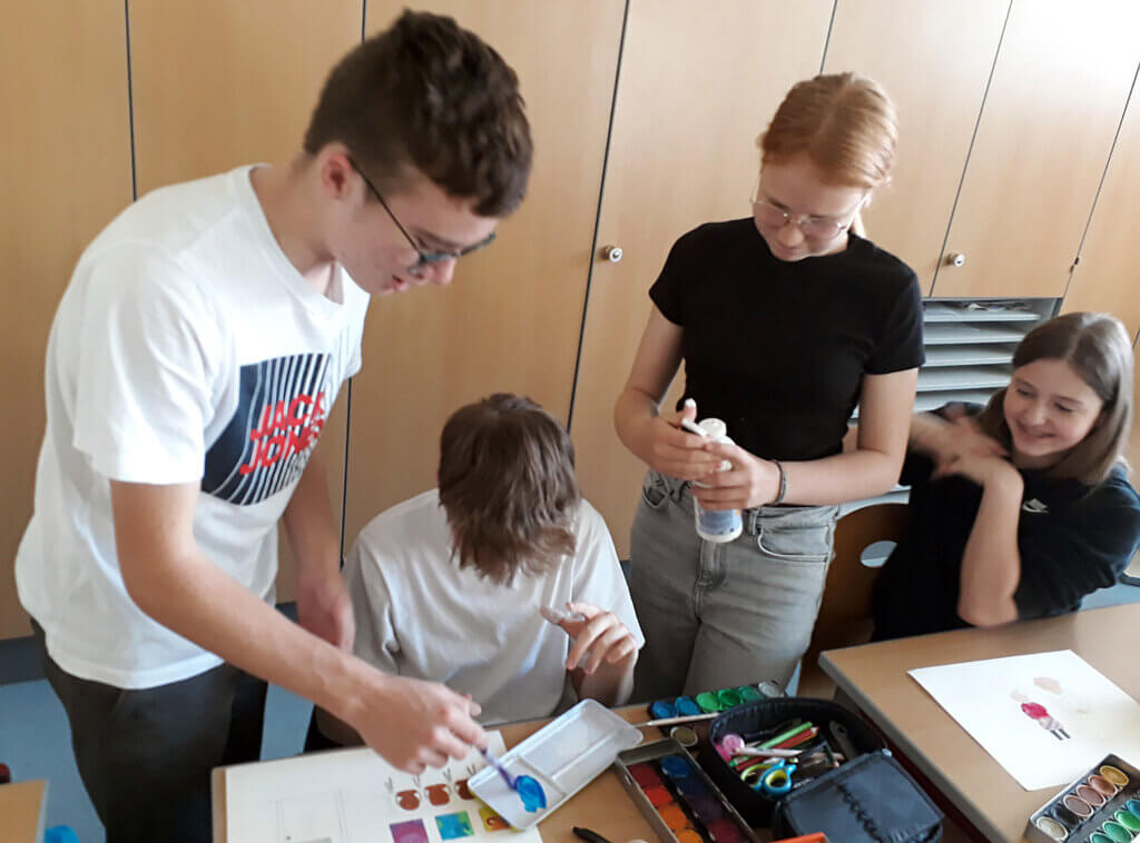 Jugendliche arbeiten im Klassenzimmer an einem kreativen Kunstprojekt mit Farben, Pinseln und Papier.