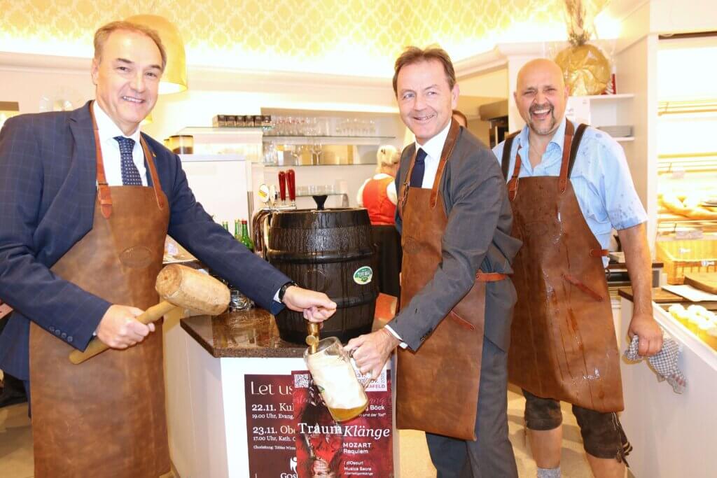 Bayer_02 Männer zapfen in traditioneller Kleidung ein Bierfass in einer Bäckerei an, Teil eines regionalen Events für Qualitätsjournalismus.