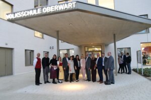 Menschen stehen vor der Volksschule Oberwart zur Eröffnung. Eine Gruppe posiert lachend für ein regionales Eventfoto.