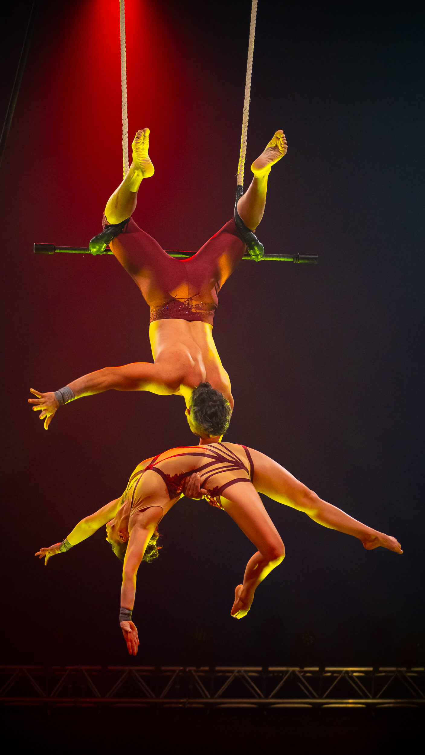 Akrobaten am Trapez in eleganten Posen, beleuchtet von rotem Scheinwerferlicht, vermitteln Spannung und Anmut.