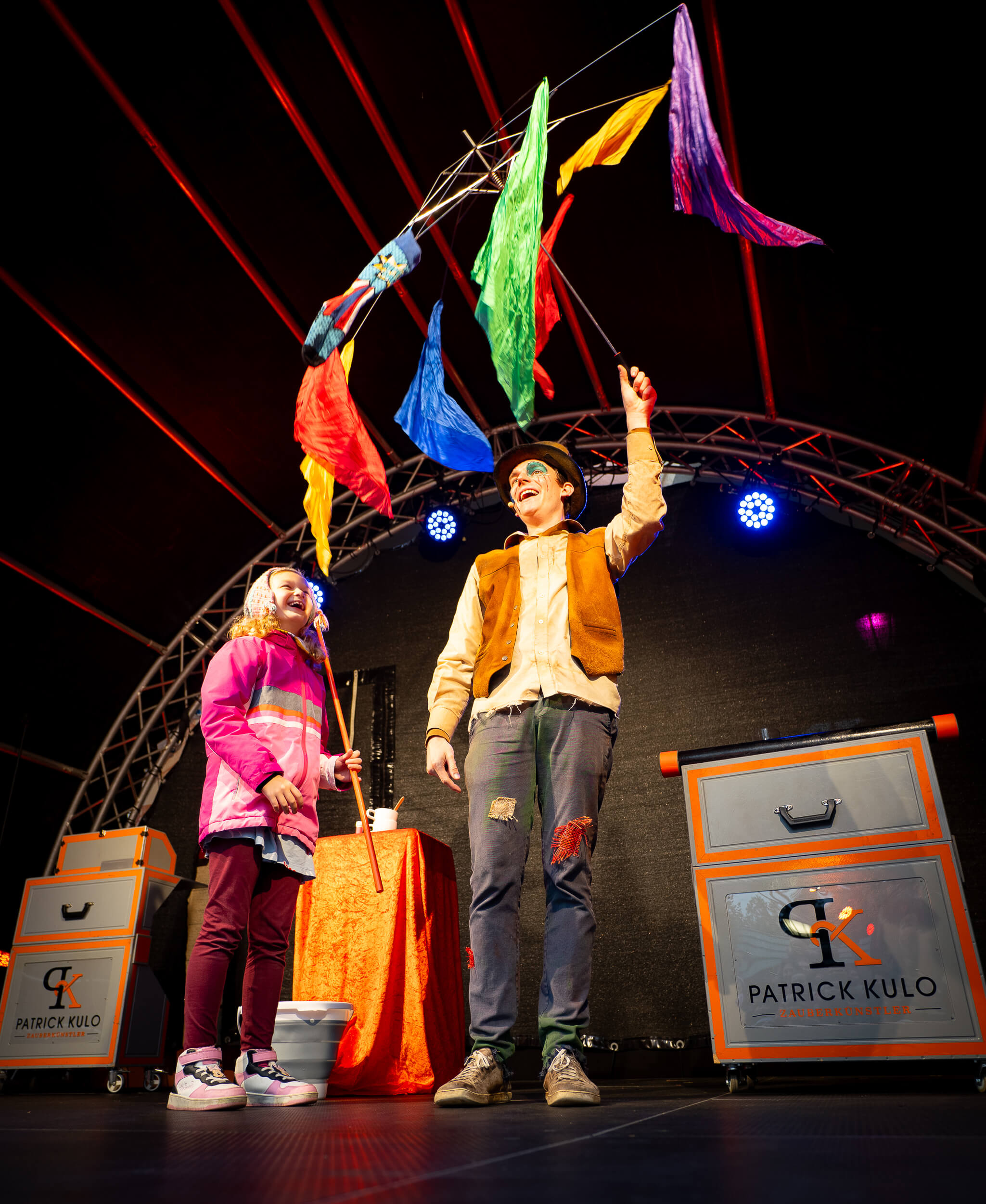 Ein fröhlicher Artist zeigt bunte Tücher auf einer Bühne einem lachenden Kind. Magischer Moment auf einer regionalen Veranstaltung.