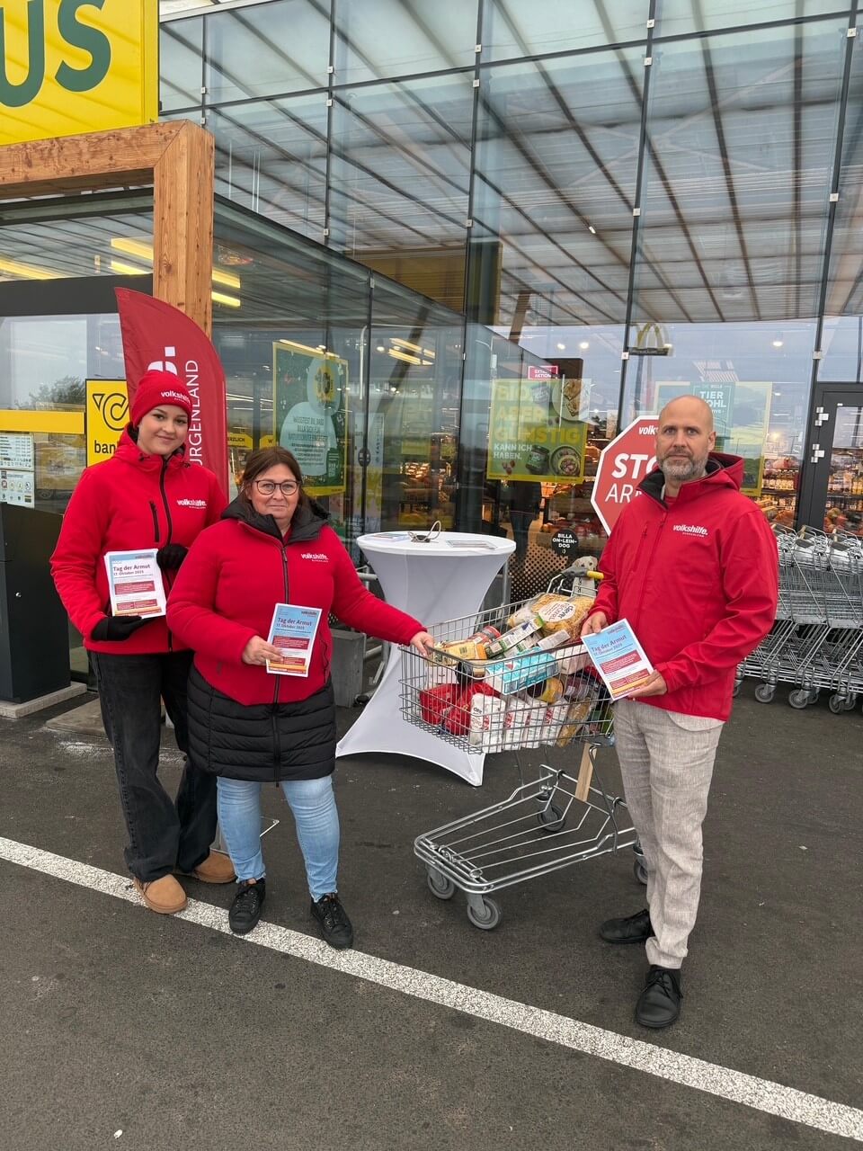 Drei Personen in roten Jacken sammeln Spenden mit einem vollen Einkaufswagen vor einem Supermarkt für einen wohltätigen Zweck.