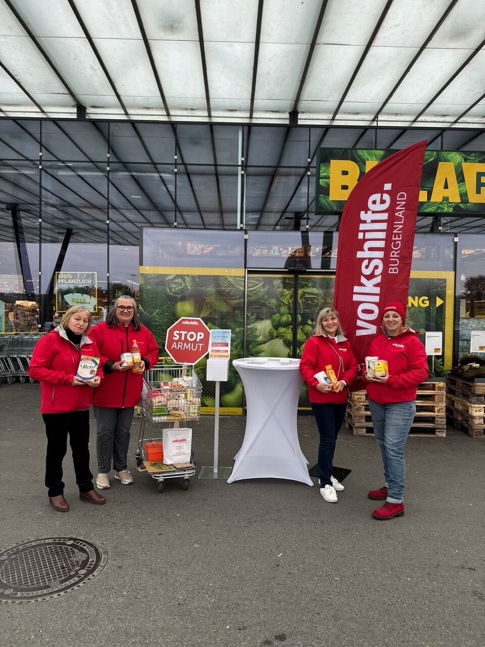 Vier Personen in roten Jacken sammeln vor einem Supermarkt Spenden für Bedürftige, unterstützt von einem lokalen Verein.