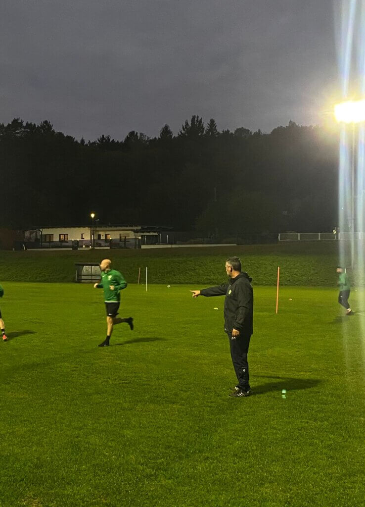 Fußballtrainer gibt Anweisungen bei abendlichem Training auf dem Sportplatz. Im Hintergrund Bäume und Gebäude.