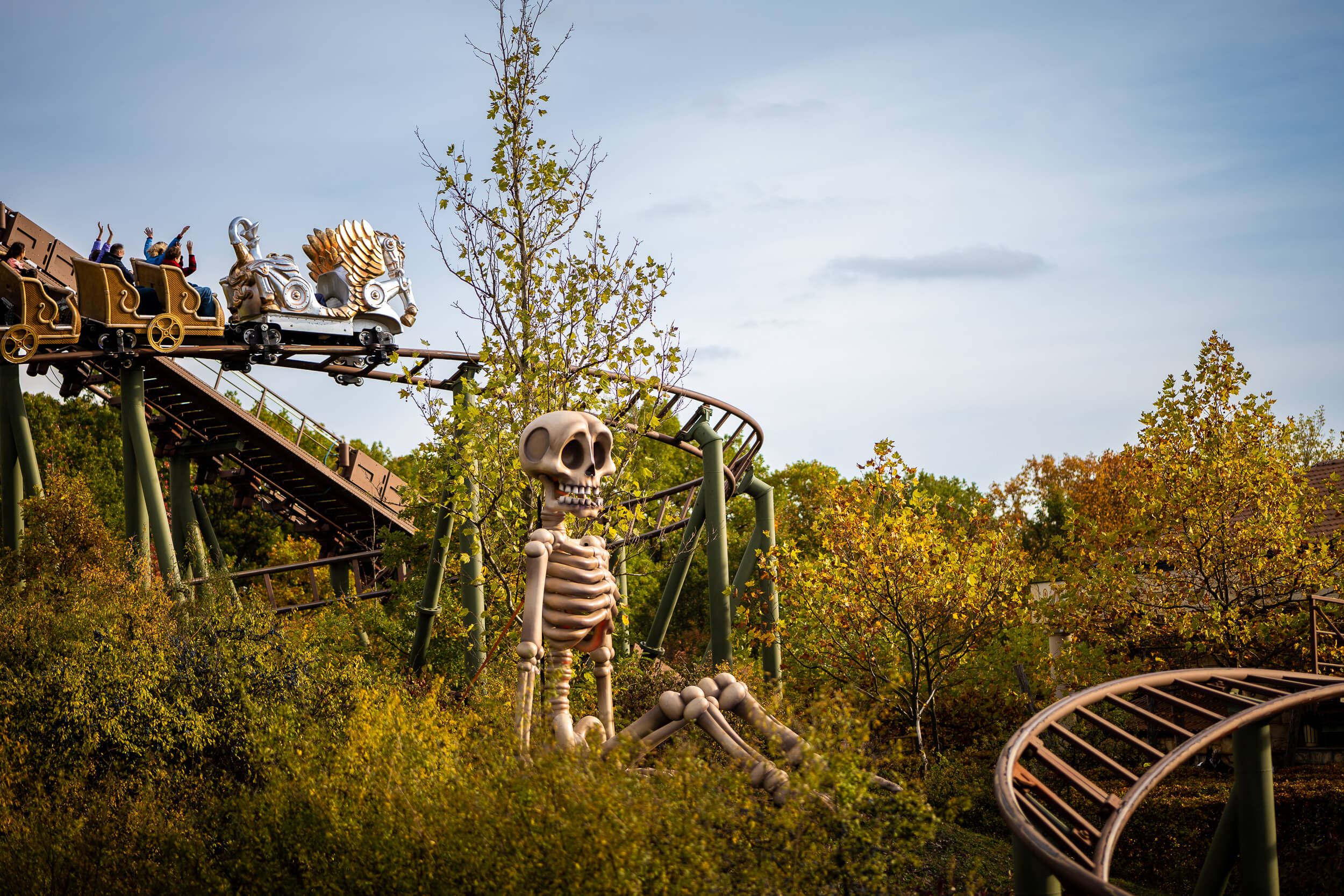 Achterbahn mit Skelett-Dekoration in herbstlicher Landschaft im Freizeitpark, Menschen heben die Arme in Freude.