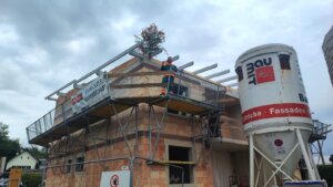 Richtfest auf einer Baustelle mit geschmücktem Baum auf dem Dach und Bauarbeiter auf einem Gerüst, im Hintergrund ein Mischer.