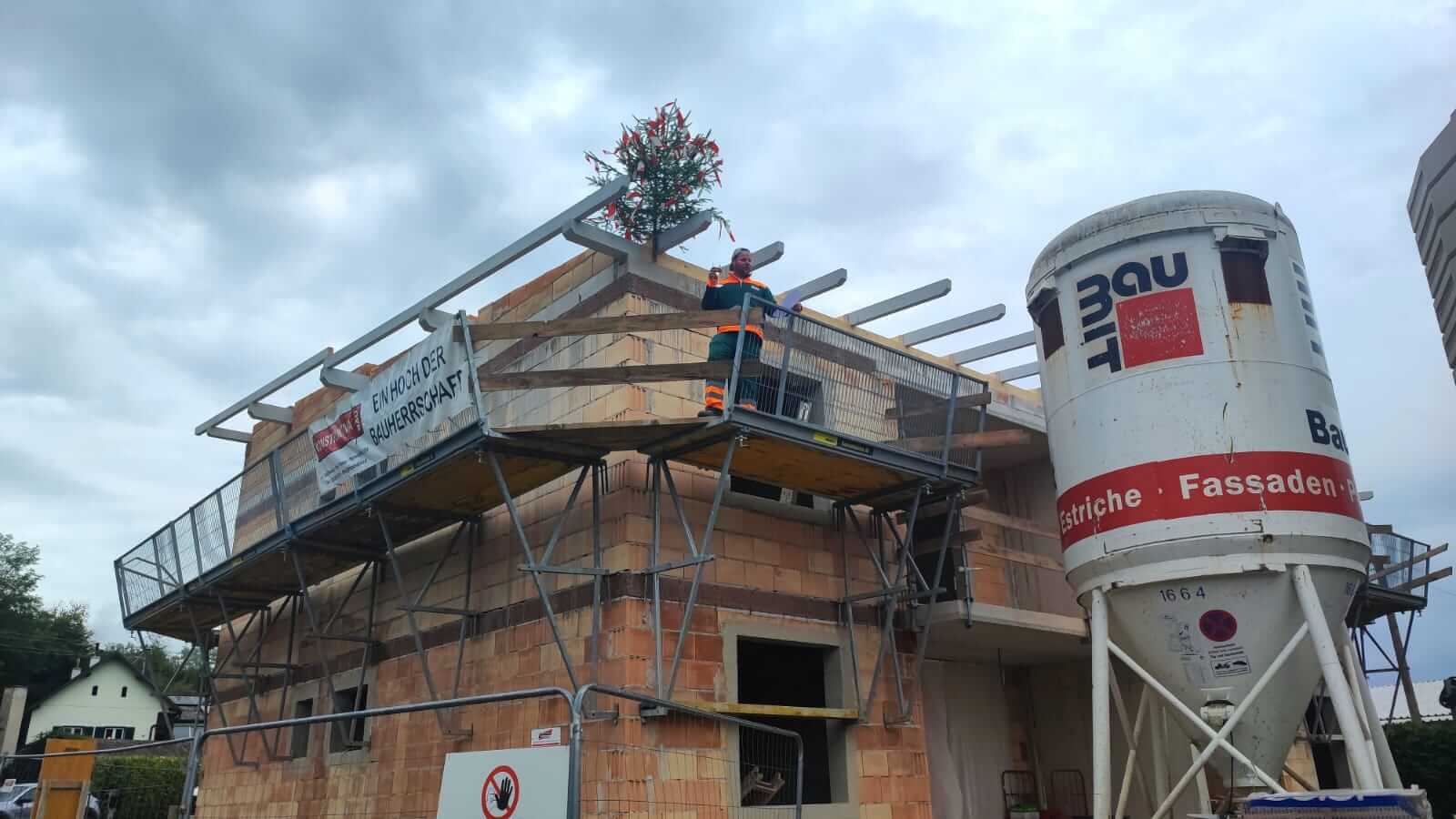 Richtfest auf einer Baustelle mit geschmücktem Baum auf dem Dach und Bauarbeiter auf einem Gerüst, im Hintergrund ein Mischer.