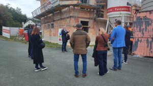 Personengruppe bei einer Besichtigung eines regionalen Bauprojekts, geführt von einem Experten, vor einem im Bau befindlichen Gebäude.