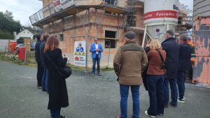 Gruppe besichtigt Baustelle eines neuen Wohnprojekts; ein Mann in blauem Blazer spricht vor dem Rohbau.