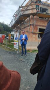 Ein Mann in blauer Jacke steht vor einer Baustelle und einem Sicherheitszaun mit Schild, während er spricht.