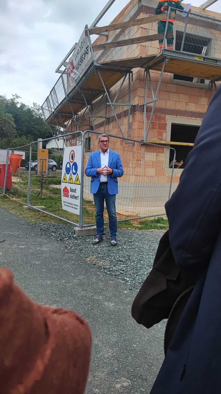 Ein Mann in blauer Jacke steht vor einer Baustelle und einem Sicherheitszaun mit Schild, während er spricht.