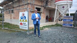 Person in blauer Jacke vor einer Baustelle, spricht neben einem Sicherheitszaun mit Hinweisschild.