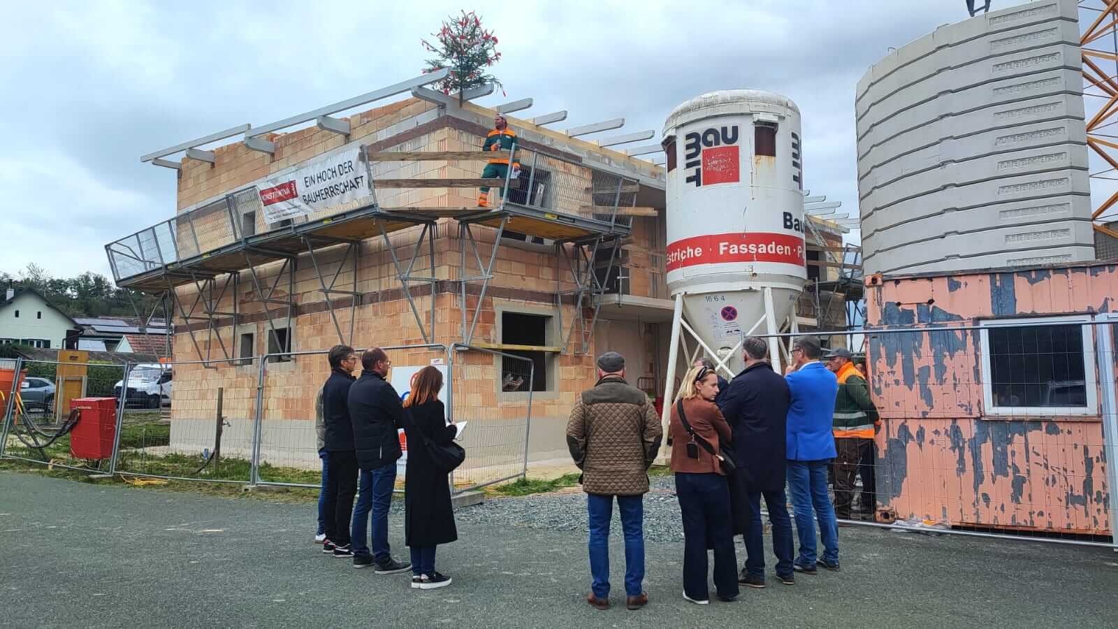 Richtfest eines Neubaus: Menschen versammeln sich vor einem Rohbau mit geschmücktem Dach, Bauarbeiter hält eine Rede.