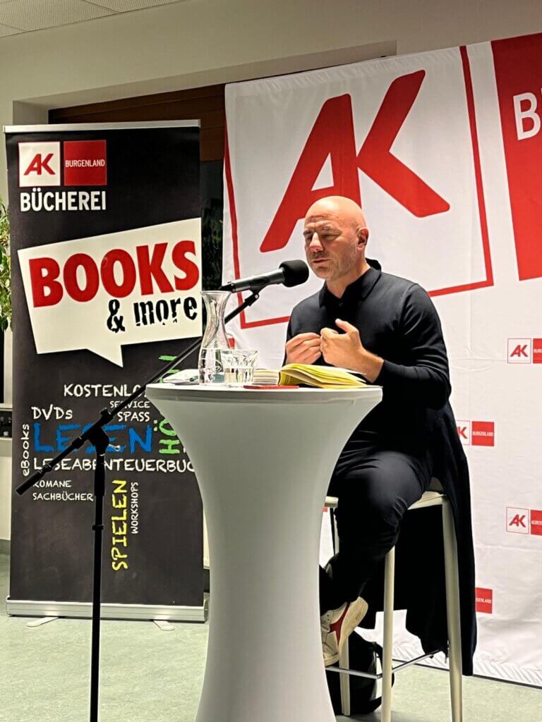 IMG-20251015-WA0002 Eine Lesung in der AK-Bücherei mit einer Person am Mikrofon. Veranstaltungsbanner im Hintergrund.