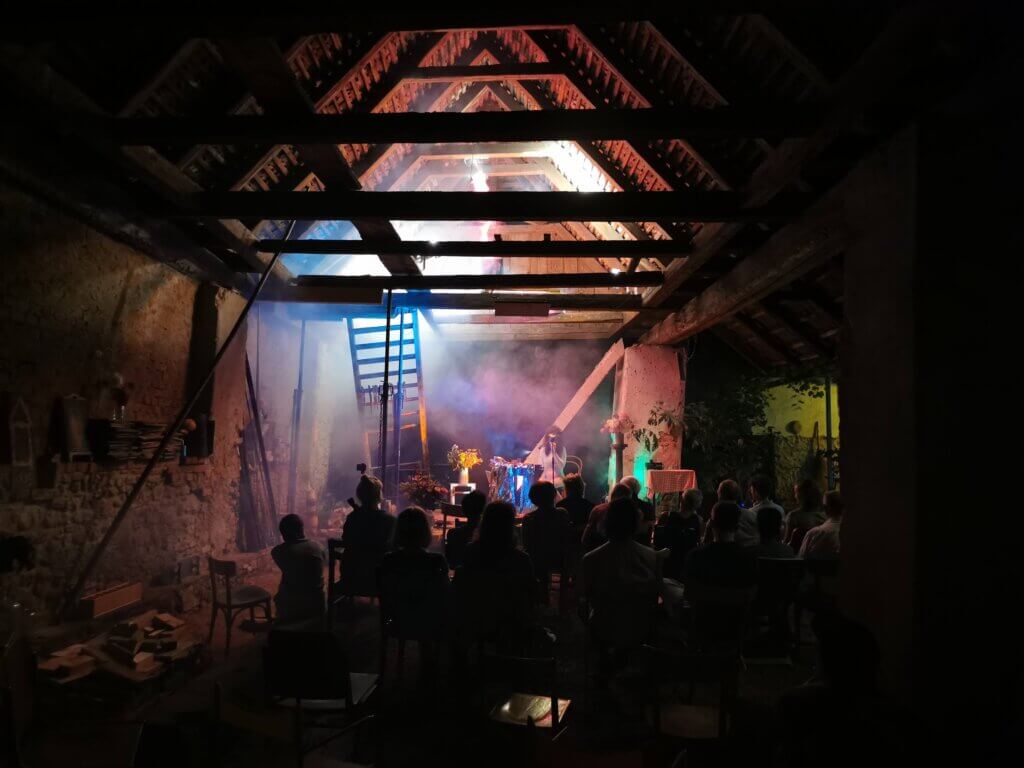 Konzert in rustikaler Scheune mit stimmungsvoller Beleuchtung, Menschen genießen Livemusik. Ideal für regionales Magazin-Feature.