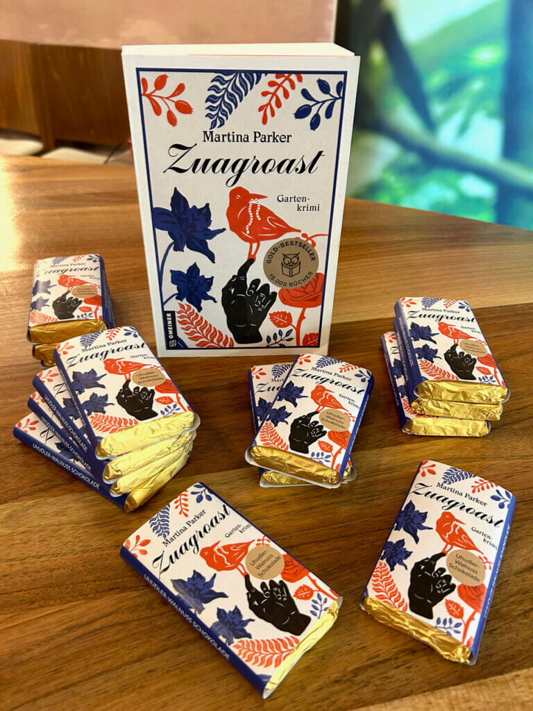 IMG_8371 Buch Zuagroast von Martina Parker mit dazu passenden Schokoladen auf Holztisch präsentiert im Prima Magazin-Kontext.