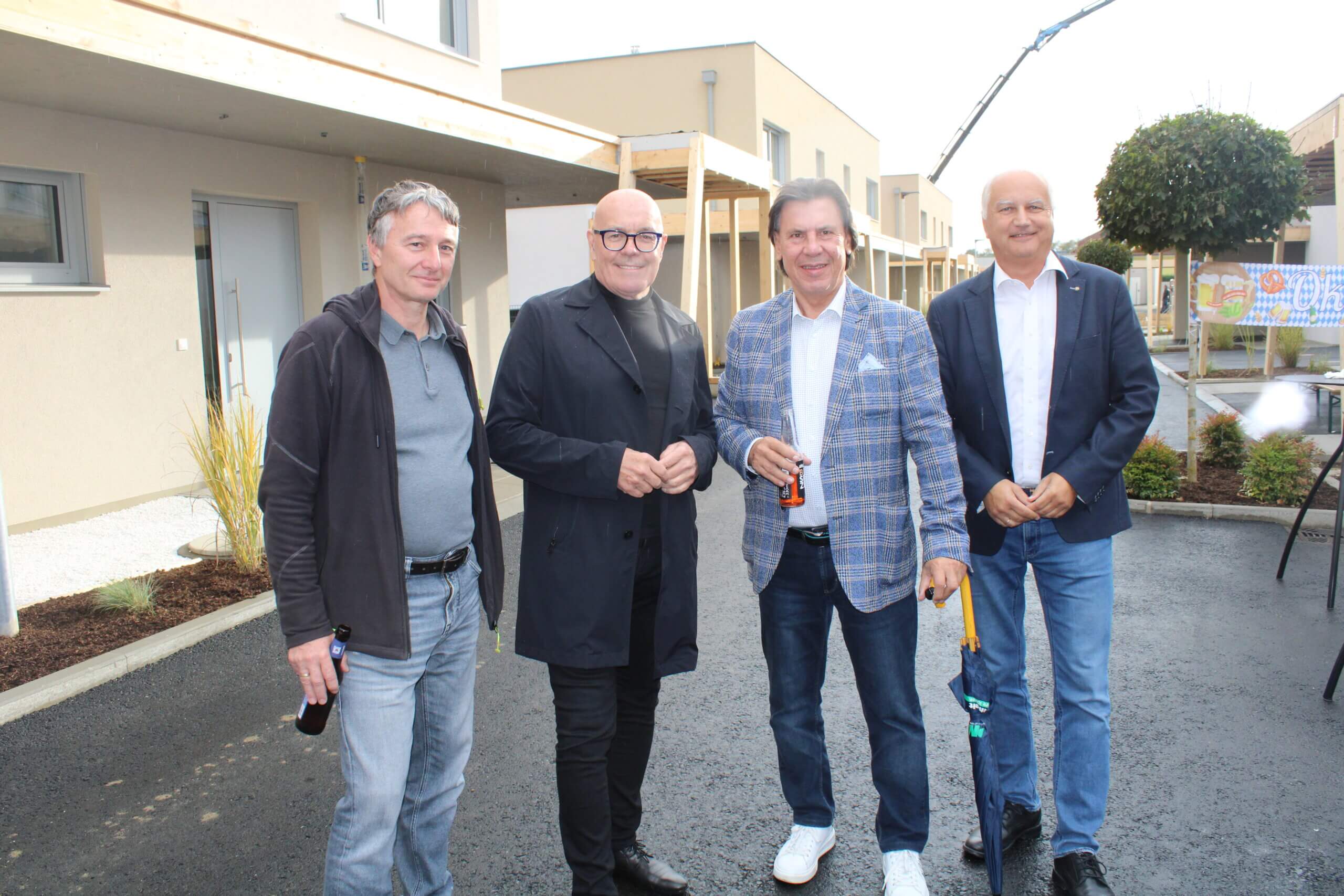 Vier Männer stehen bei der Eröffnung eines modernen Wohnprojekts auf einer gepflasterten Fläche vor moderner Architektur.