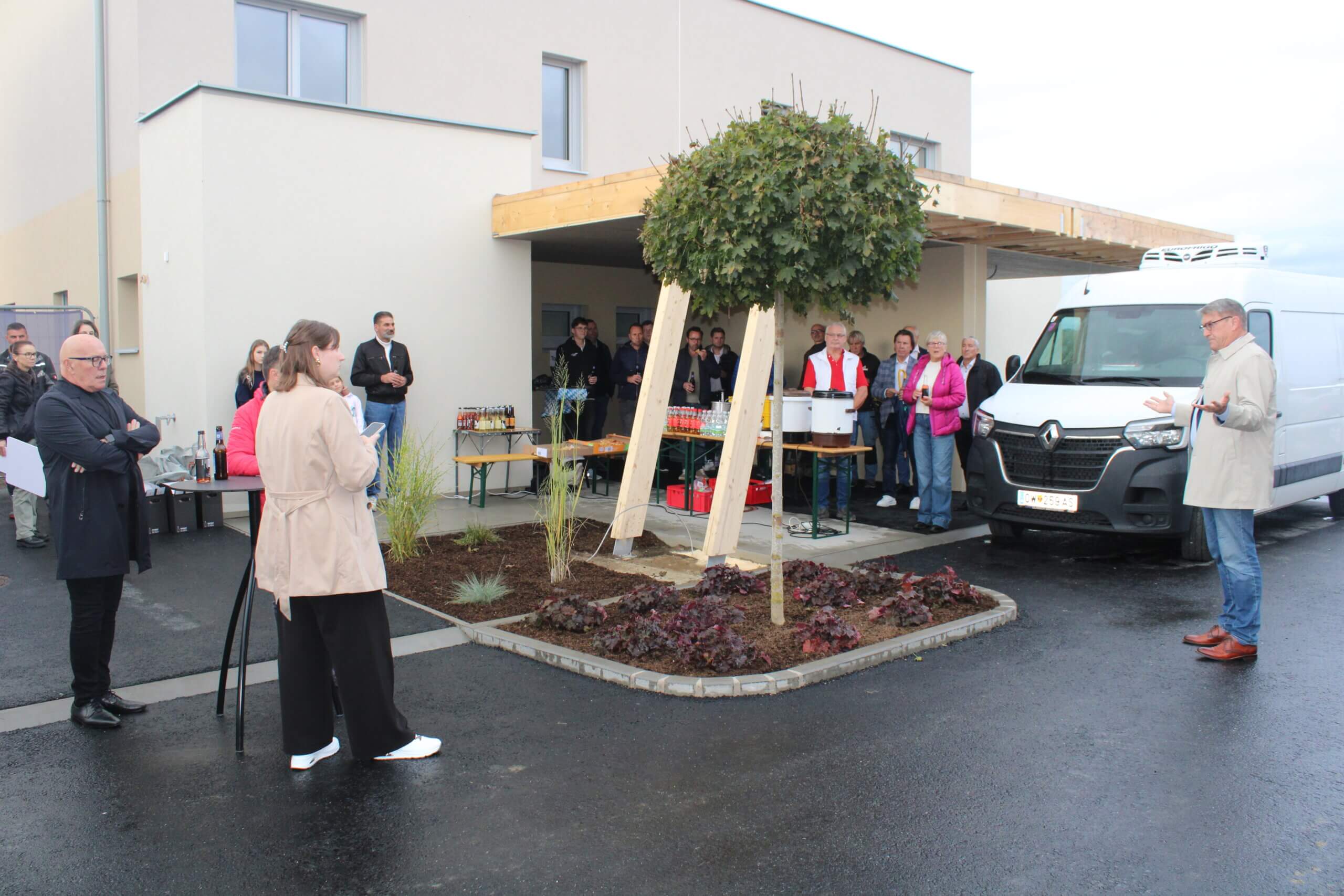 Menschen versammeln sich vor einem modernen Gebäude zu einem Event, während ein Mann spricht. Getränke und ein Lieferwagen im Hintergrund.