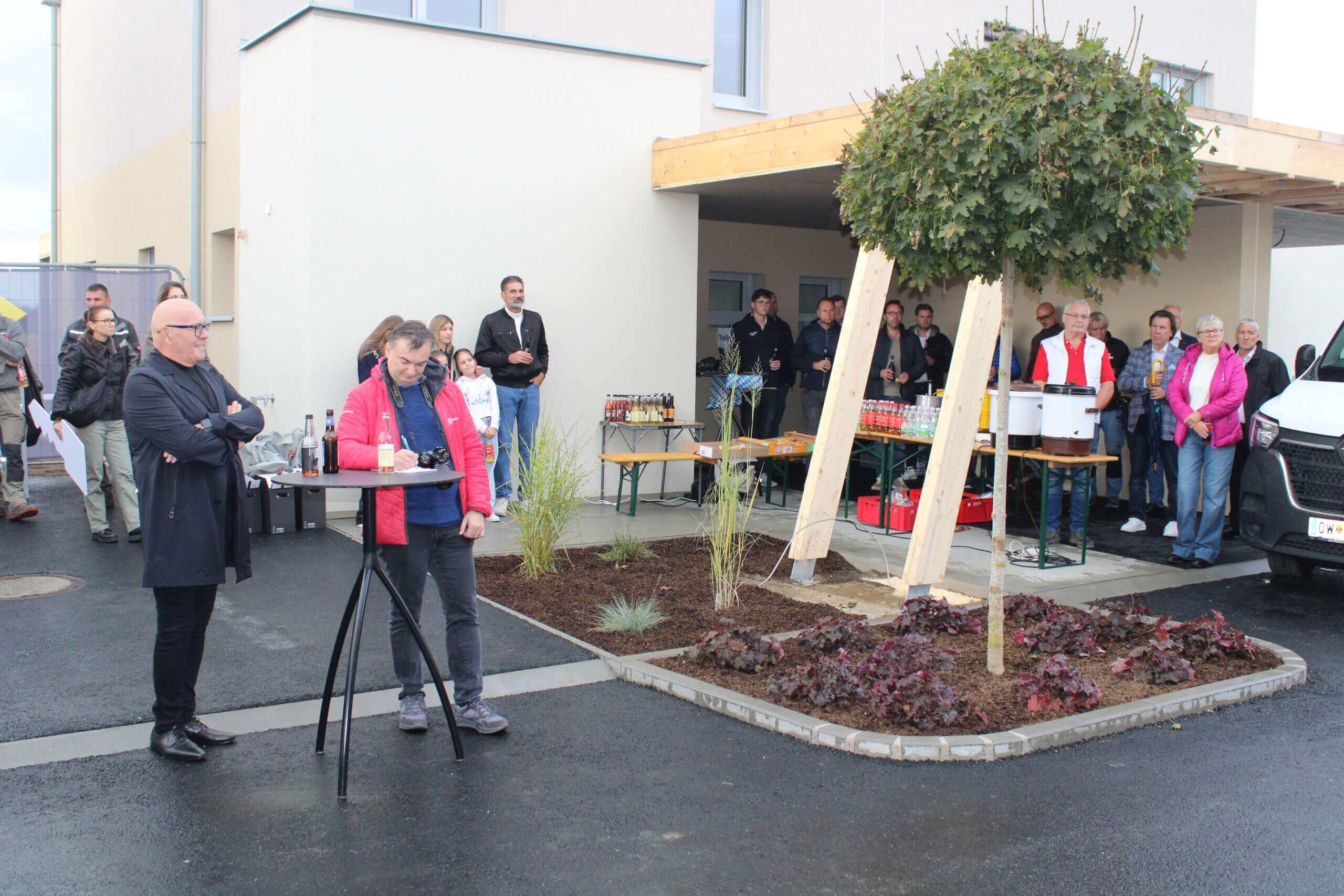 Menschen bei einer Feier im Freien vor einem modernen Gebäude, mit Getränken und Snacks auf Tischen unter einem Baum.