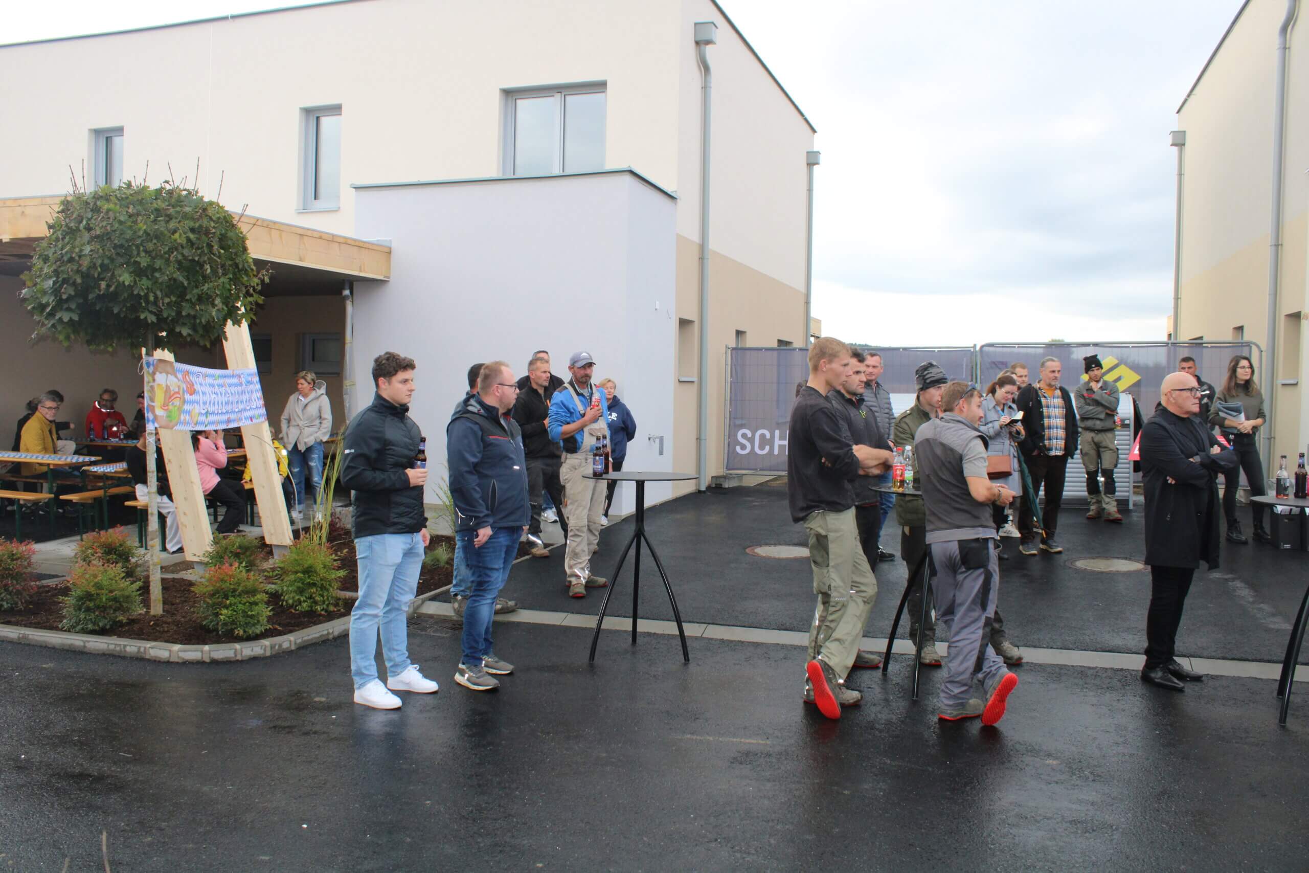 Menschen bei einer Veranstaltung im Freien vor modernen Gebäuden, einige halten Getränke. Geselliges Beisammensein einer Gemeinschaft.