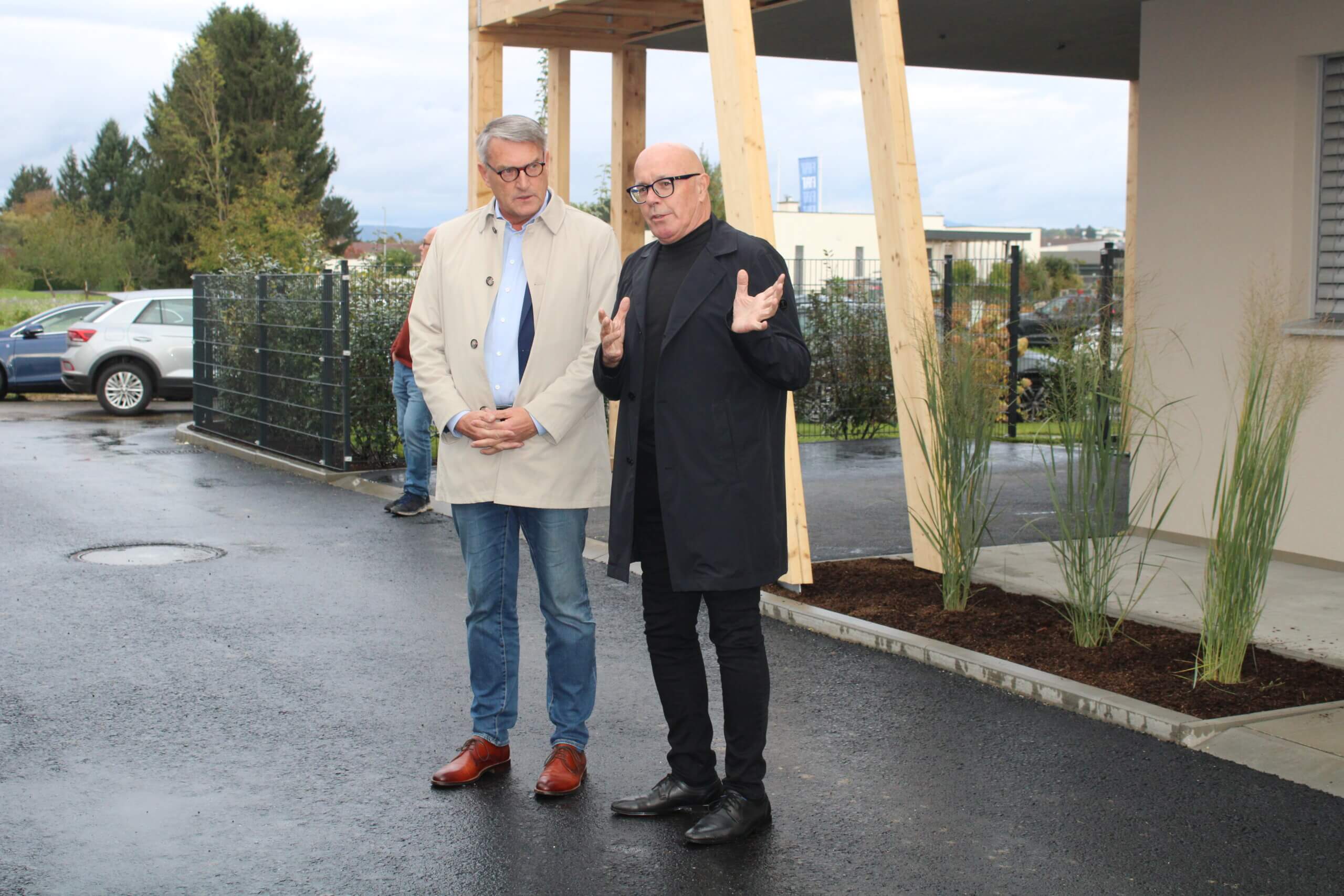 Zwei Männer diskutieren im Freien vor einem modernen Gebäude, umgeben von Pflanzen, für einen Artikel im Prima Magazin.