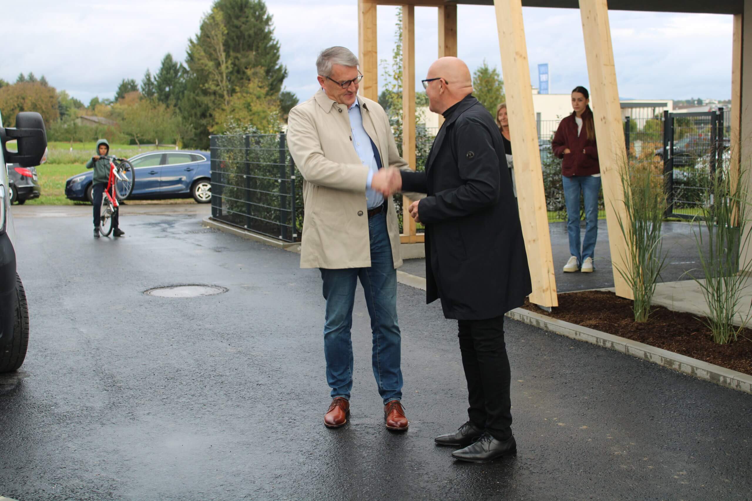 Zwei Männer schütteln sich die Hände vor einem Gebäude, während ein Junge mit Fahrrad vorbeigeht.