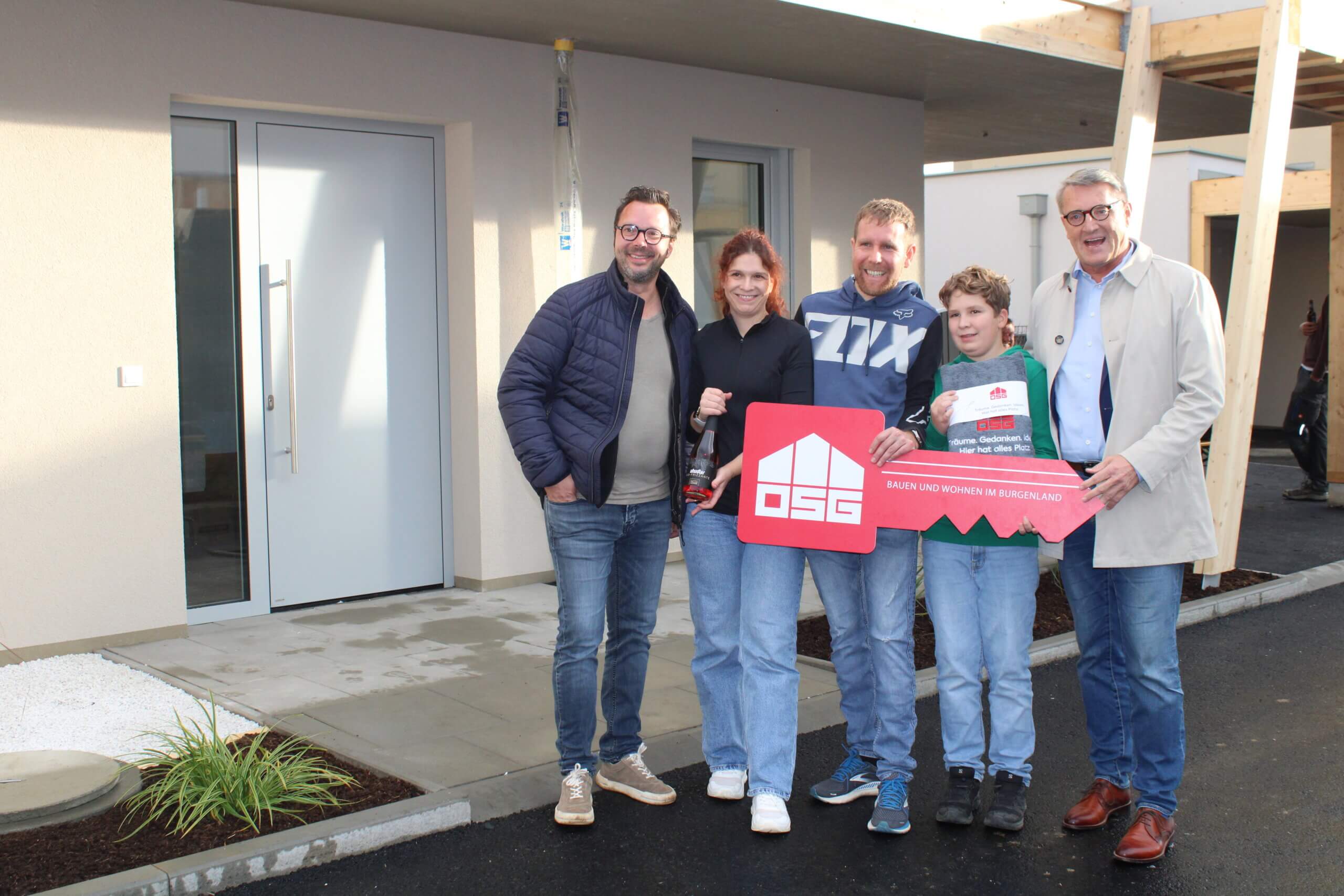 Familie posiert vor neuem Haus mit symbolischem Schlüssel und Sektflasche, feierlich im Burgenland, unterstützt von OSG.