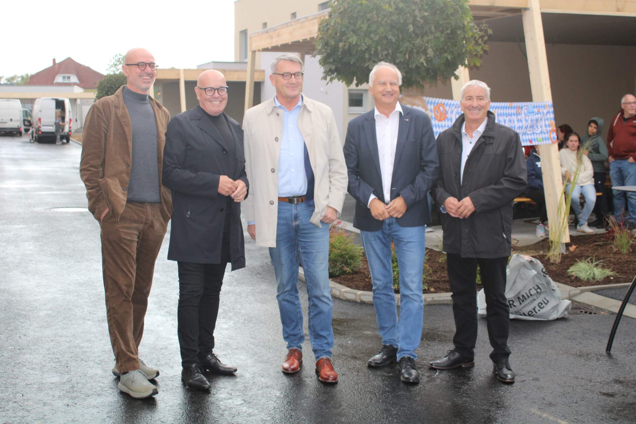 Fünf Männer posieren lächelnd bei einer Veranstaltung im Freien, während im Hintergrund Gäste auf einer überdachten Terrasse sitzen.