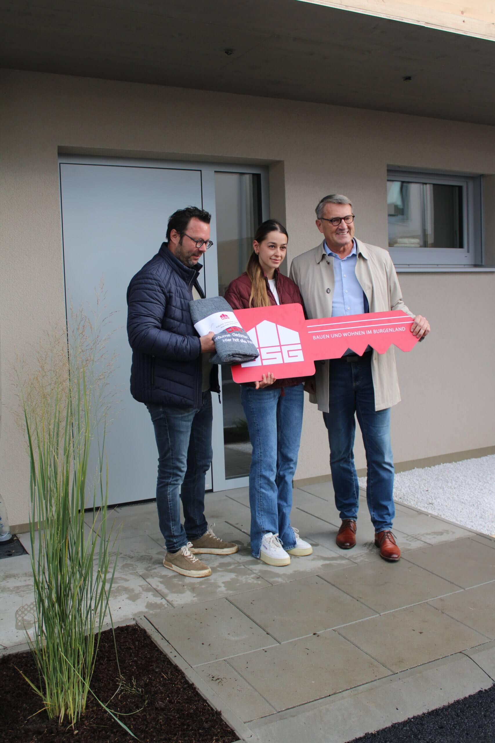 Menschen posieren lächelnd mit symbolischem Schlüssel vor ihrem neuen Zuhause, übergeben von einem Bauträger.